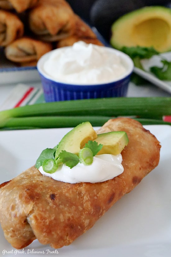 A white plate of fried burrito with sour cream, sliced ​​avocado, scallions and coriander on top.