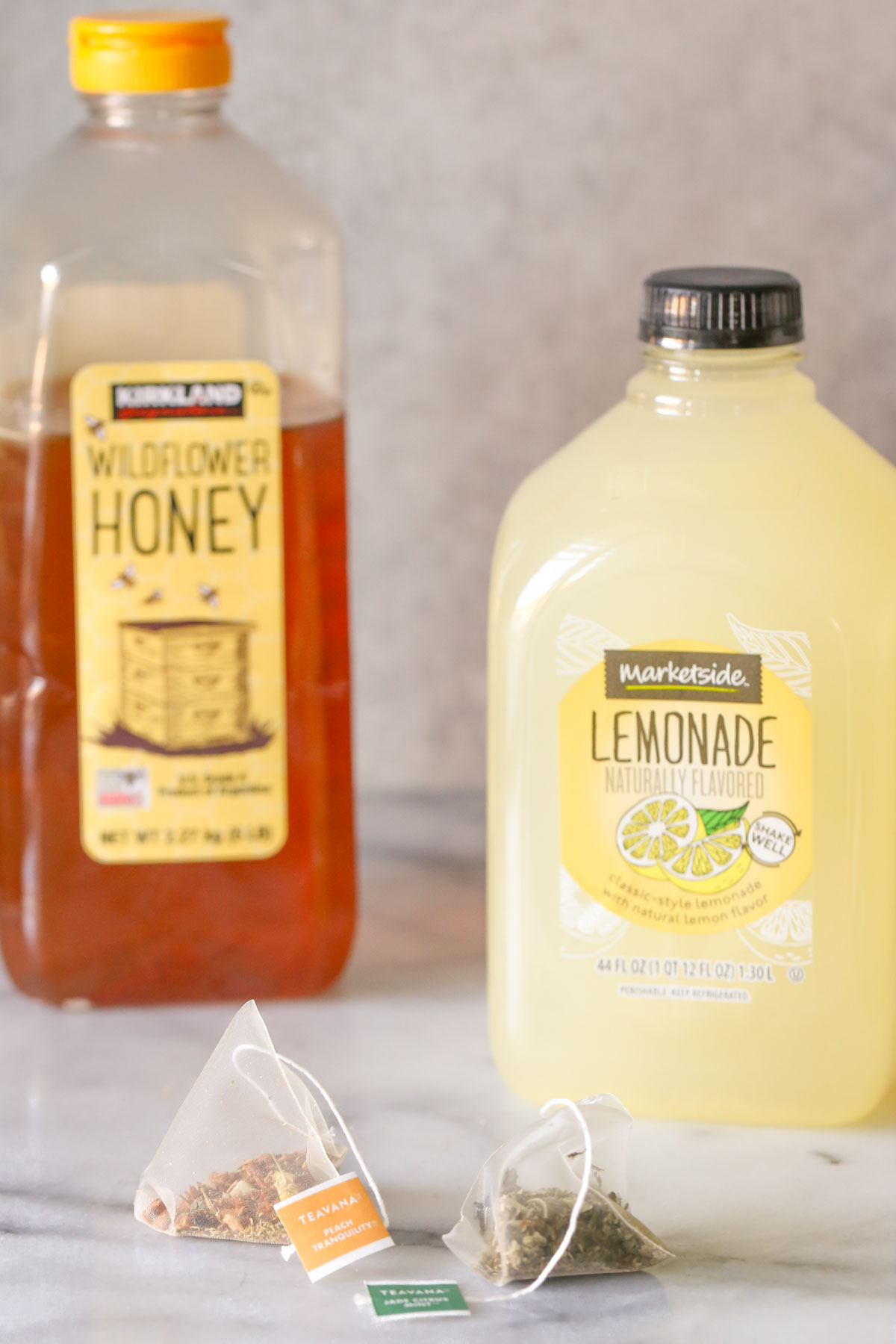 A pack of Teavana Peach Tranquility tea next to a pack of Teavana Jade Citrus Mint, with a pitcher of lemonade and a container of honey in the back, all the ingredients for the Starbucks Medicine Ball Tea.