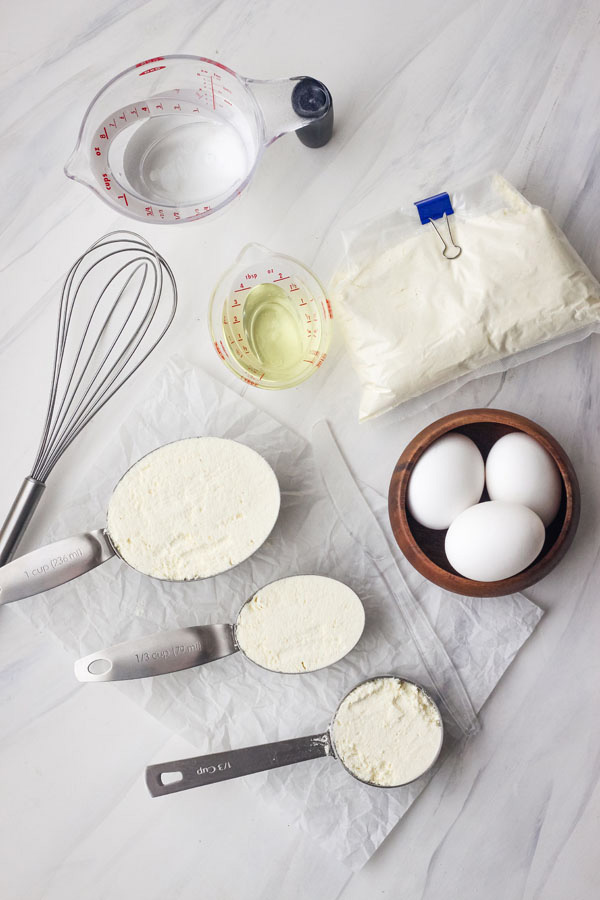 A white table with measuring cups, a whisk and eggs.