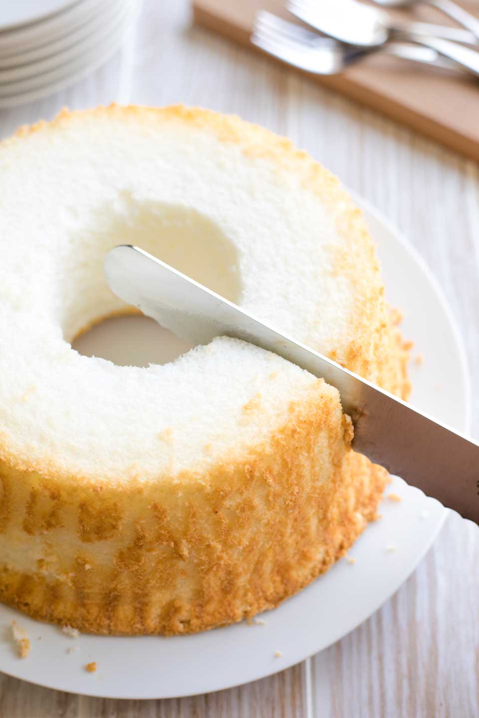 The white plate holds an entire angel cake, with a long serrated knife just beginning to make the first cut.