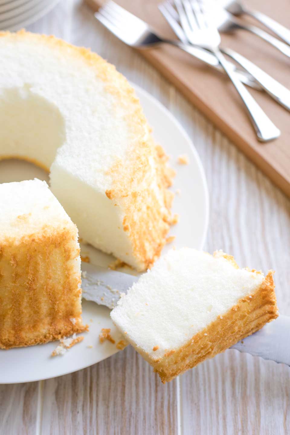 A slice of angel food cake, placed on the serrated knife that cut it, on a white plate containing the rest of the uncut pie. Forks and a stack of plates in the back.