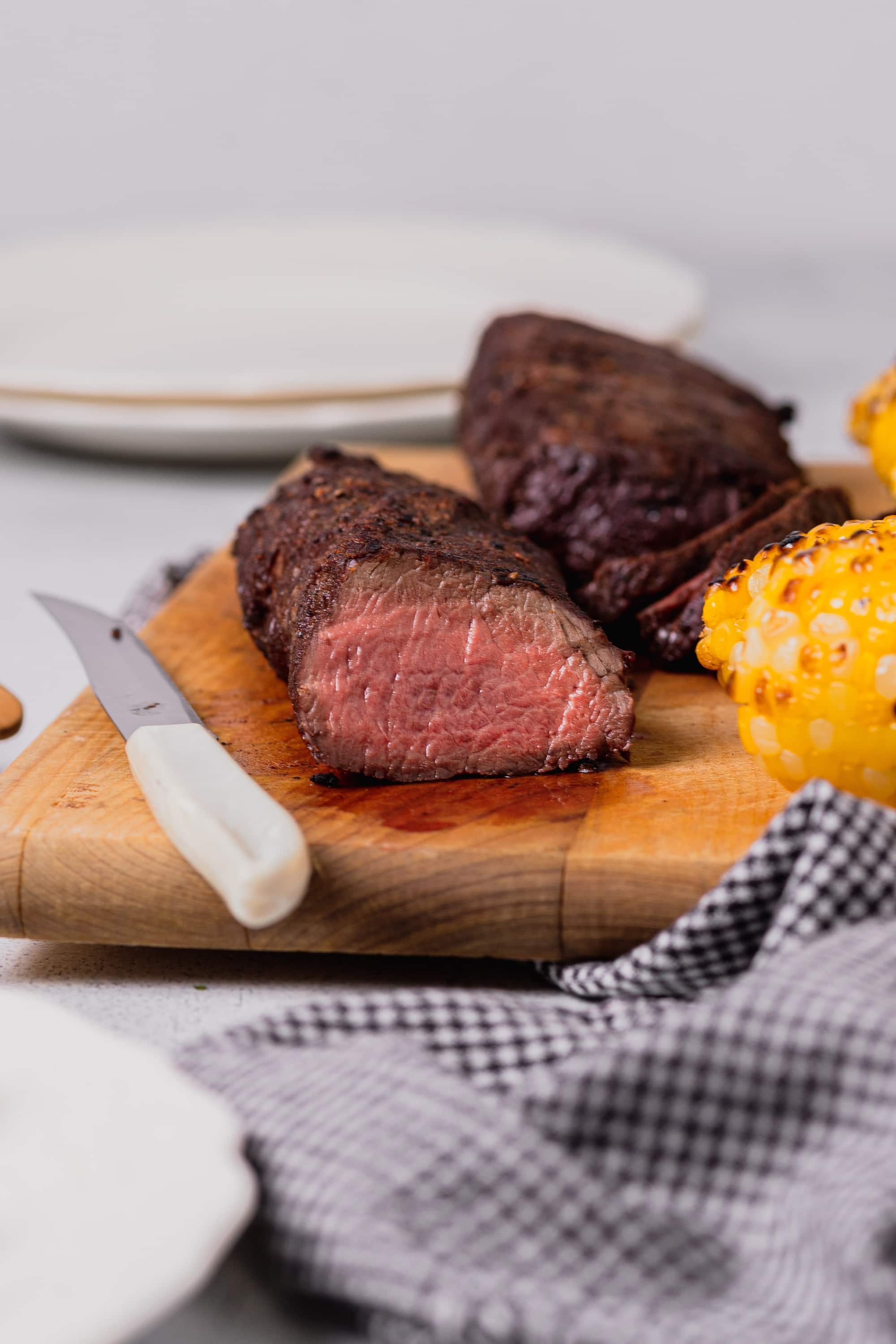 A straight shot of the roast venison's belt has been cut open to reveal the center. The venison is on a wooden cutting board along with the grilled corn.