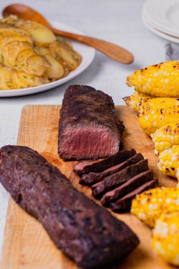 Grilled venison slices are presented on a white plate with roasted corn on the cob and baked potatoes.