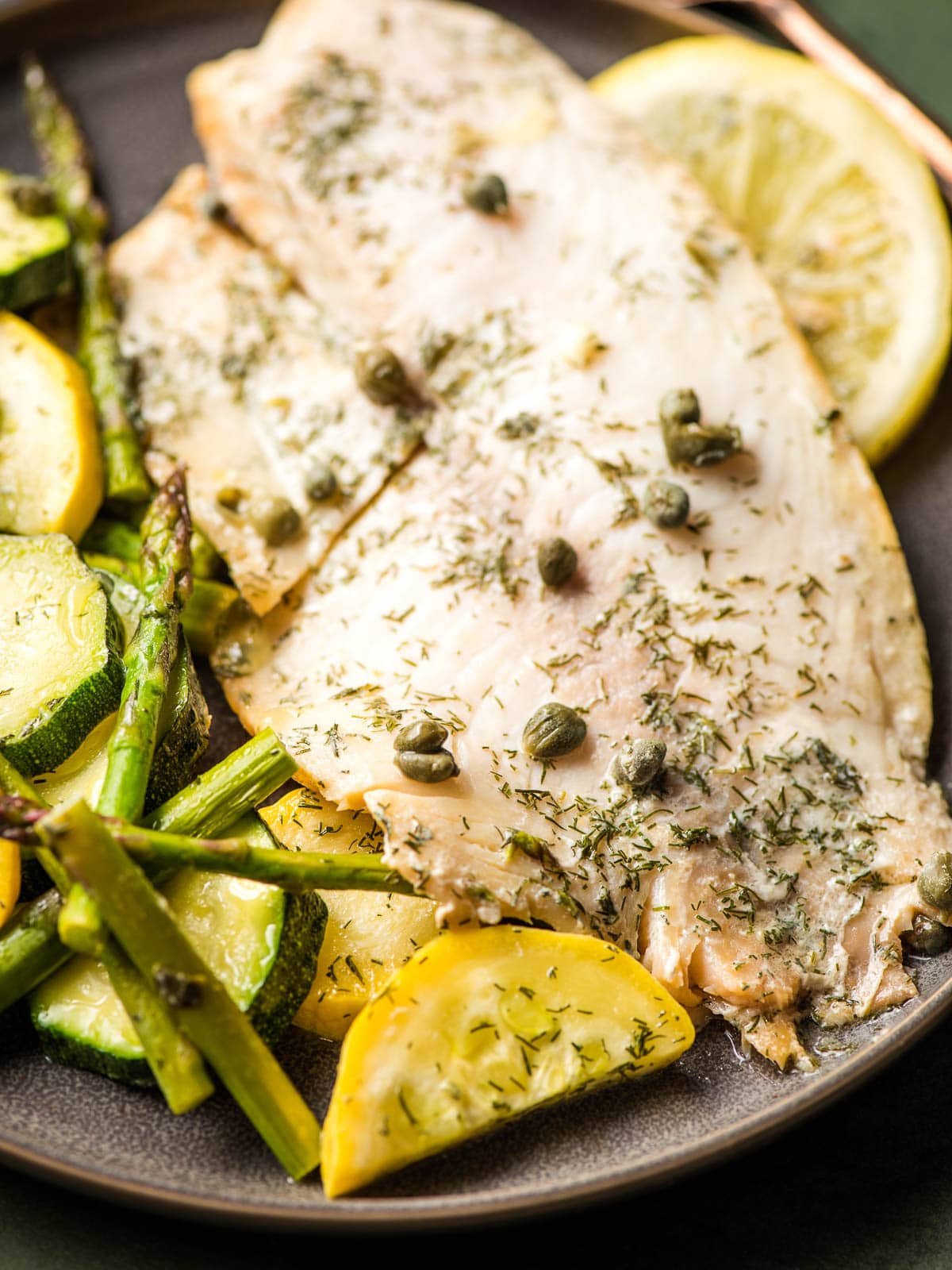 Spiced tilapia fillet with capers on a plate with sliced ​​squash and asparagus.