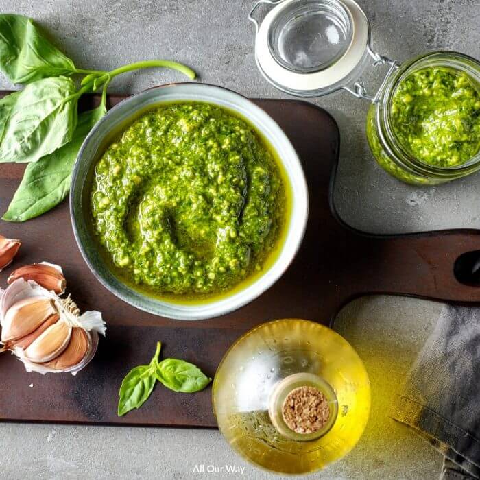 Green basil in a bowl with olive oil and garlic cloves on a wooden cutting board.