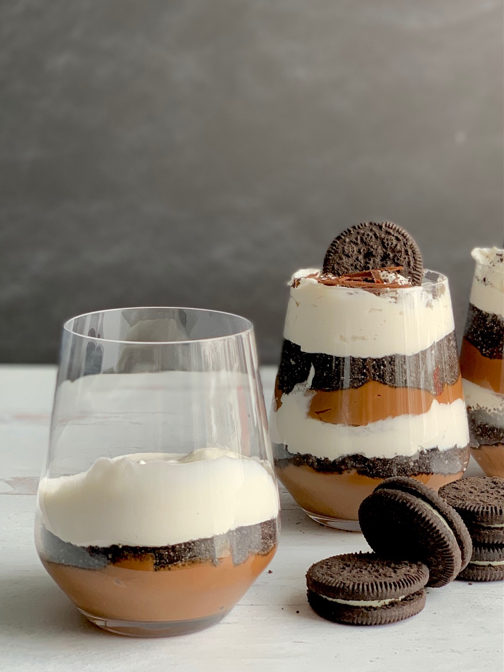 View from above 3 glass jars layered diagonally with homemade thick chocolate pudding, crushed oreo-style cookies and creamy white whipped topping. Then, topped with crushed biscuits, an oreo-like cookie, and chocolate chips. Next to the glass jars are cookies more like Oreo.