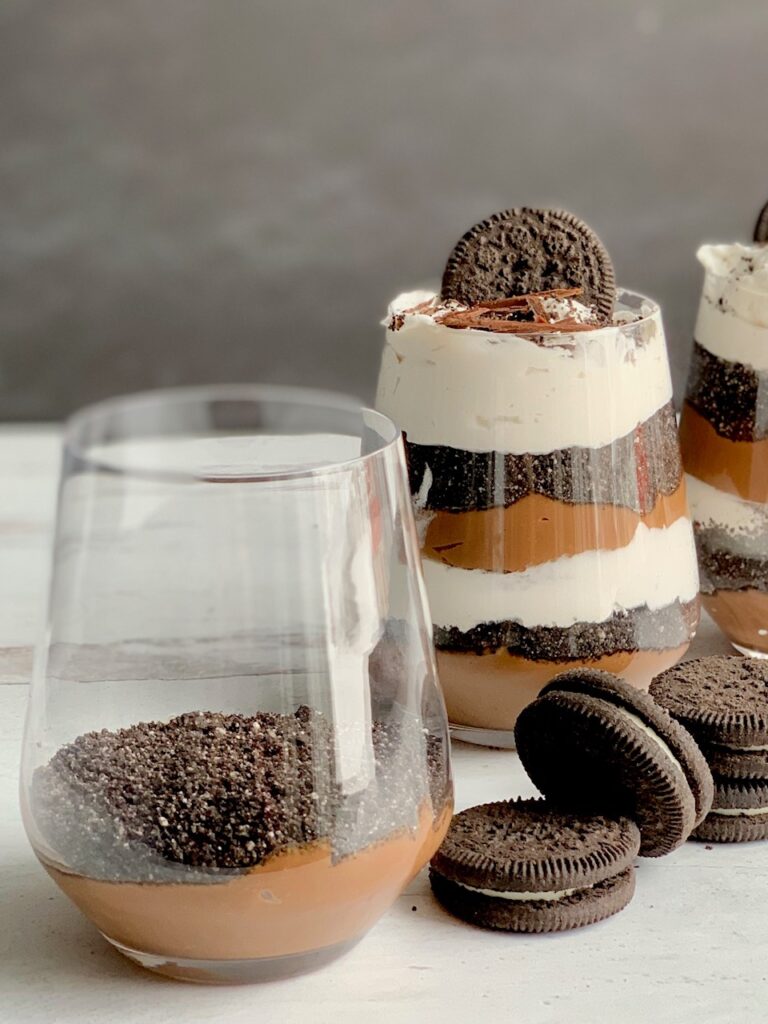3 glass jars layered diagonally with homemade thick chocolate pudding, crushed oreo cookies, and white cream topping. Then, topped with crushed biscuits, an oreo-like cookie, and chocolate chips. Next to the glass jars are cookies more like Oreo.
