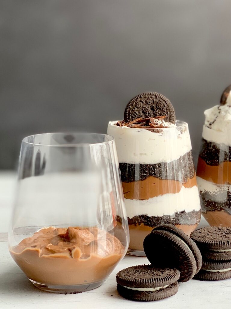 1 glass jar with the first thick layer of chocolate pudding on the bottom, then cookie crumbs and toppings next to 2 glass jars layered with homemade thick chocolate pudding, crushed oreo-style cookies and portion white cream topping. Then, topped with crushed biscuits, an oreo-like cookie, and chocolate chips. Next to the glass jars are cookies more like Oreo.