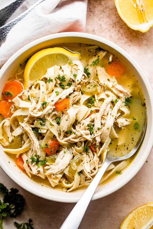 Roasted chicken noodles on high served in soup bowl with lemon and spoon inside the bowl