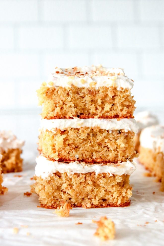 This is a stack of three pieces of cake lying on a sheet of white parchment. Many pieces of cake are blurred in the background and a white brick background is behind the stack of cakes.