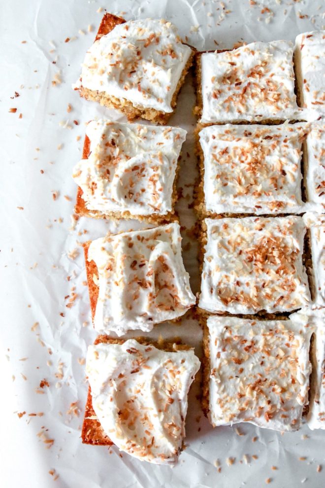 This is an overhead image of a square cut coconut cake. The cake rests on a piece of white parchment and is separated and pulled from the other pieces. The cake is covered with a layer of white cream and toasted coconut.