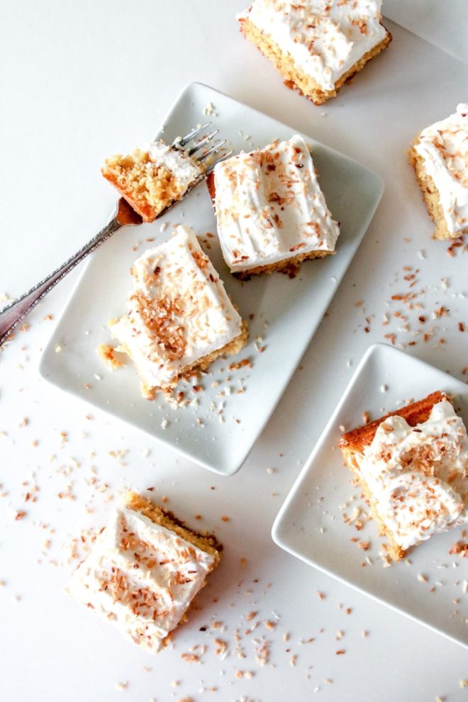 This is an overhead view of square slices of coconut cake topped with vanilla ice cream and toasted coconut. Some pieces of cake sit on small white rectangular plates while others sit on white counters.