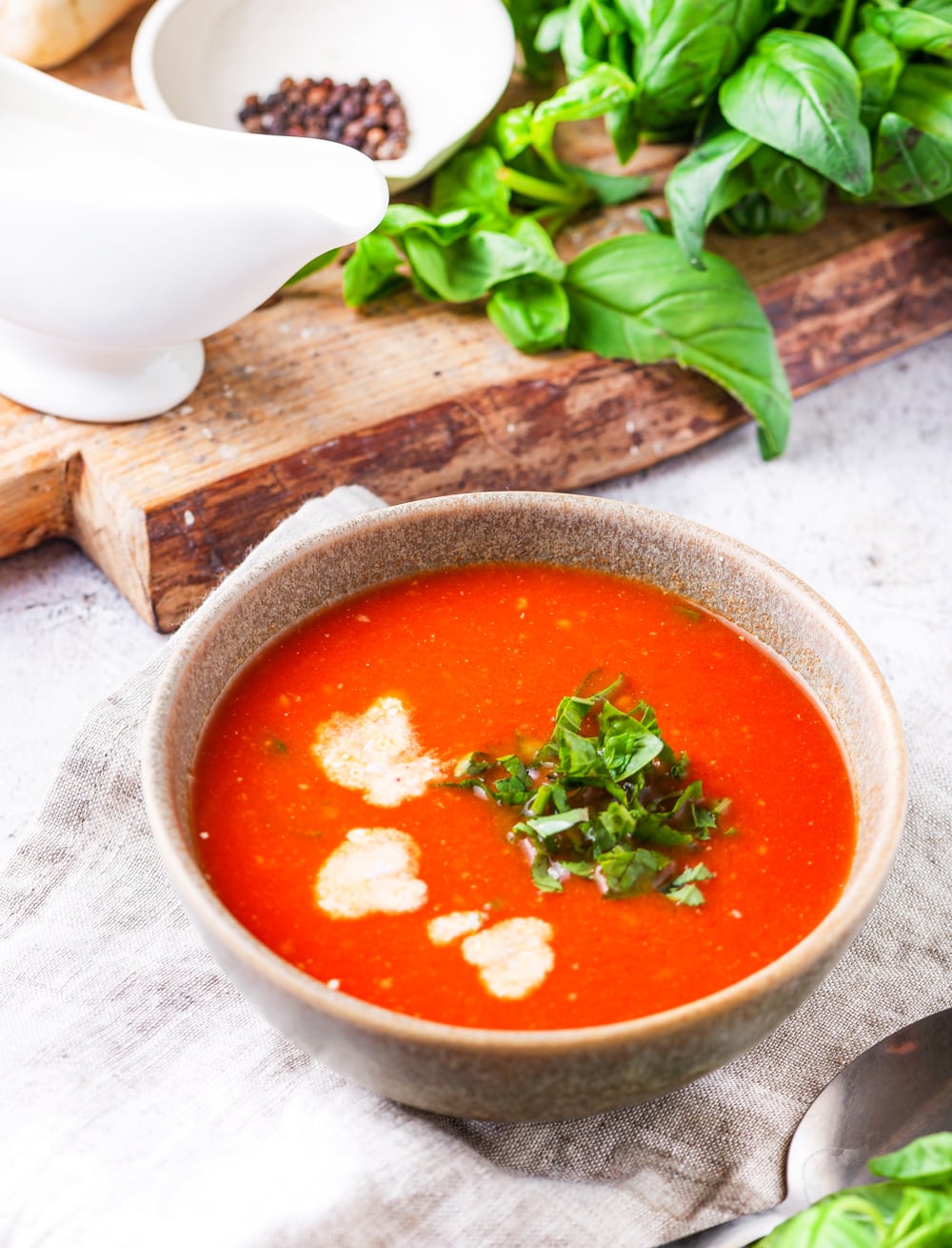 A bowl of tomato soup with a cutting board topped with basil and black pepper in the back.