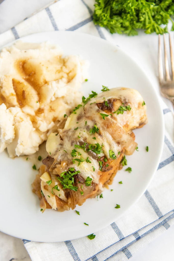 Chicken with melted cheese and onions on a white plate with mashed potatoes.