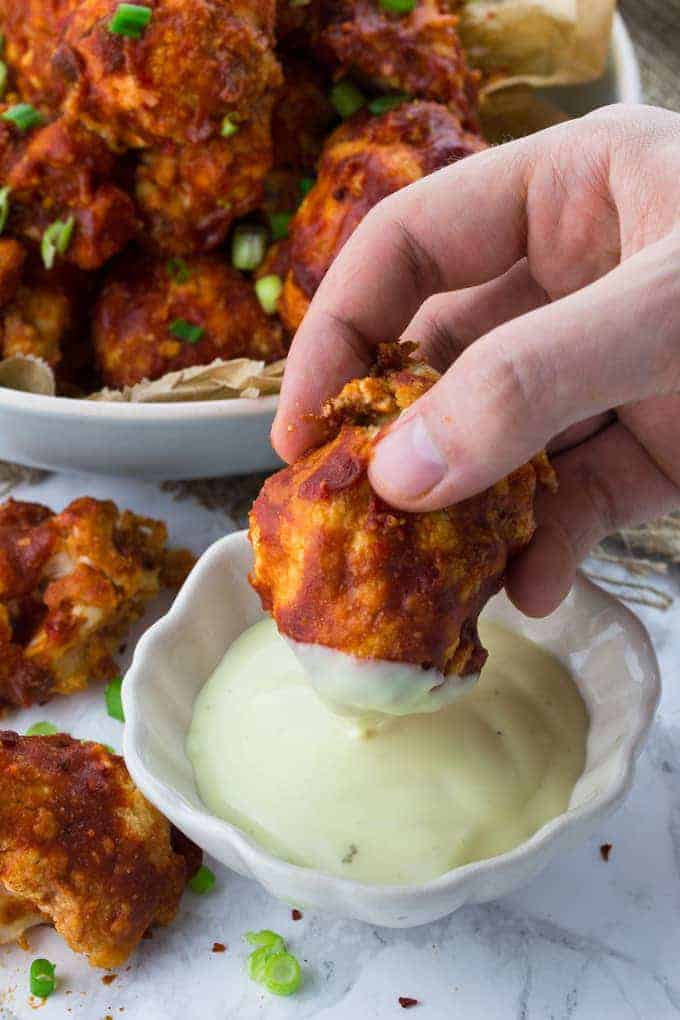 a hand dipping cauliflower wings in a small bowl of aioli with many cauliflower wings on the background