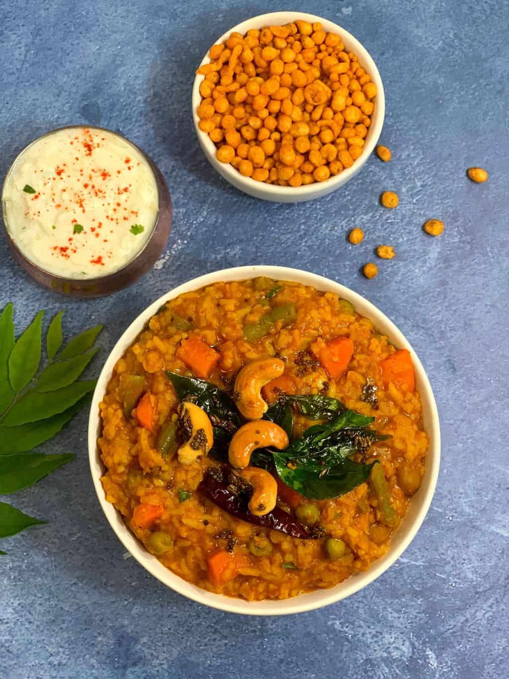 The bisi bele bath is served in a bowl with brew on top and raita and boondi on the side