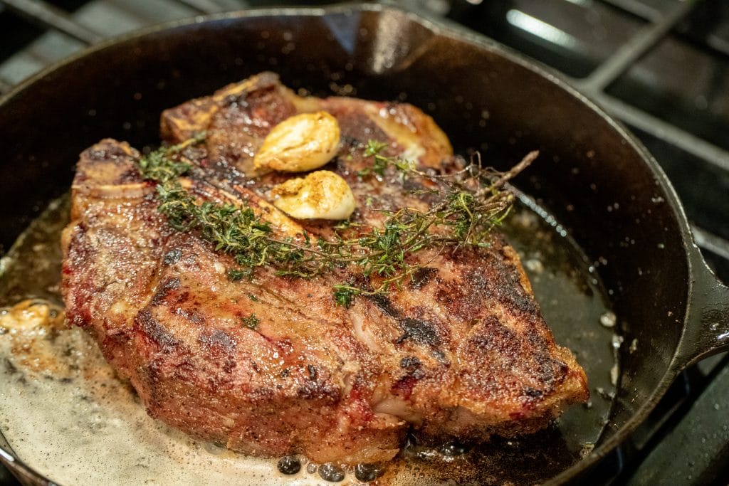 Porterhouse steak sizzling in a cast iron skillet with butter, oil, garlic cloves, and thyme sprigs.