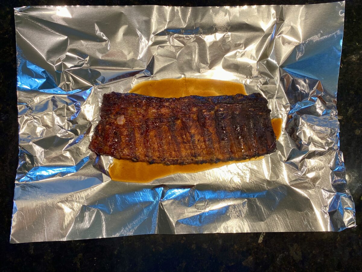 Overhead view of partially cooked rack of ribs placed meat side down on the tinfoil that has the wrap juice.