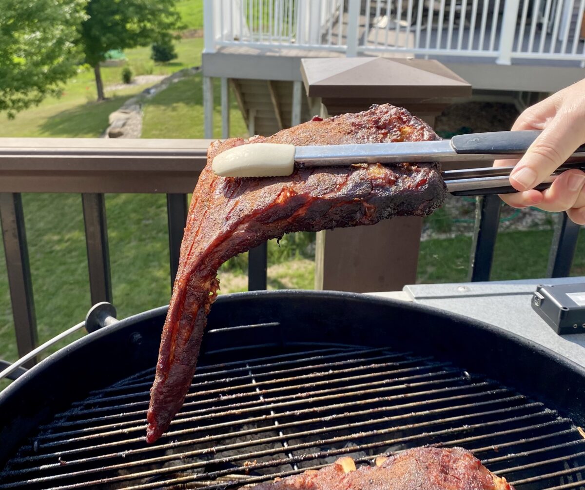 Side view of ribs being picked up with a set of tongs on the grill to show how done they are. These ribs bent to almost 90 degrees so they are almost done.