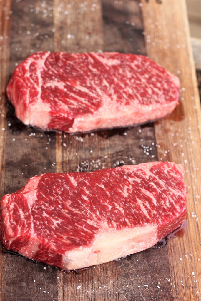 two uncooked New York strip steaks on a wooden cutting board sprinkled with salt.