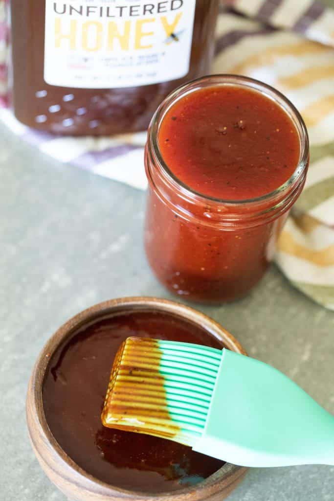Mint broom dipped in honey barbecue sauce placed over a small wooden bowl filled with honey barbecue sauce with a small mason jar filled with more sauce with a jar of honey in the back.
