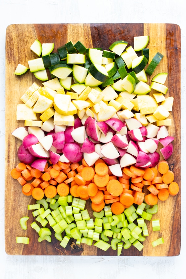 Chop zucchini, yellow squash, red potatoes, carrots, and celery on a wooden cutting board.