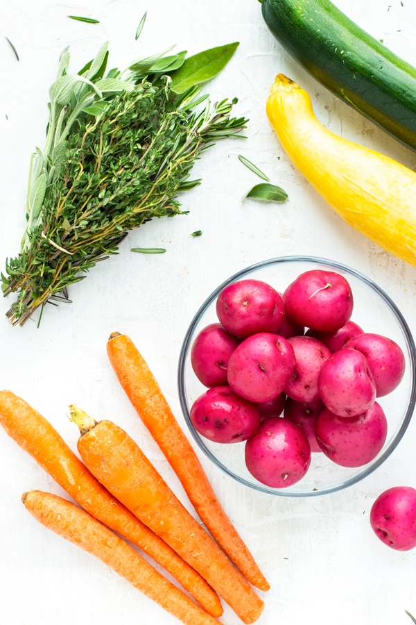 Fresh herbs, red potatoes, zucchini, yellow squash and carrots on a white background for vegetable chicken soup recipe.