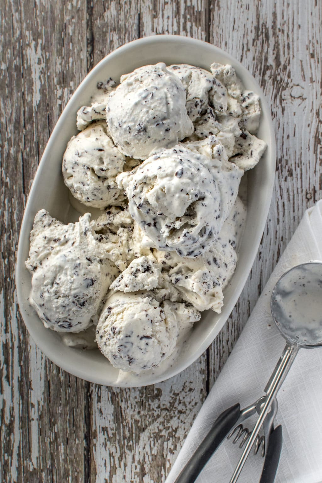 Oval, white bowl filled with vanilla, cream filled with grated chocolate. On a rustic barn board, drop back with a folded napkin on the side to place an empty scoop of ice cream.