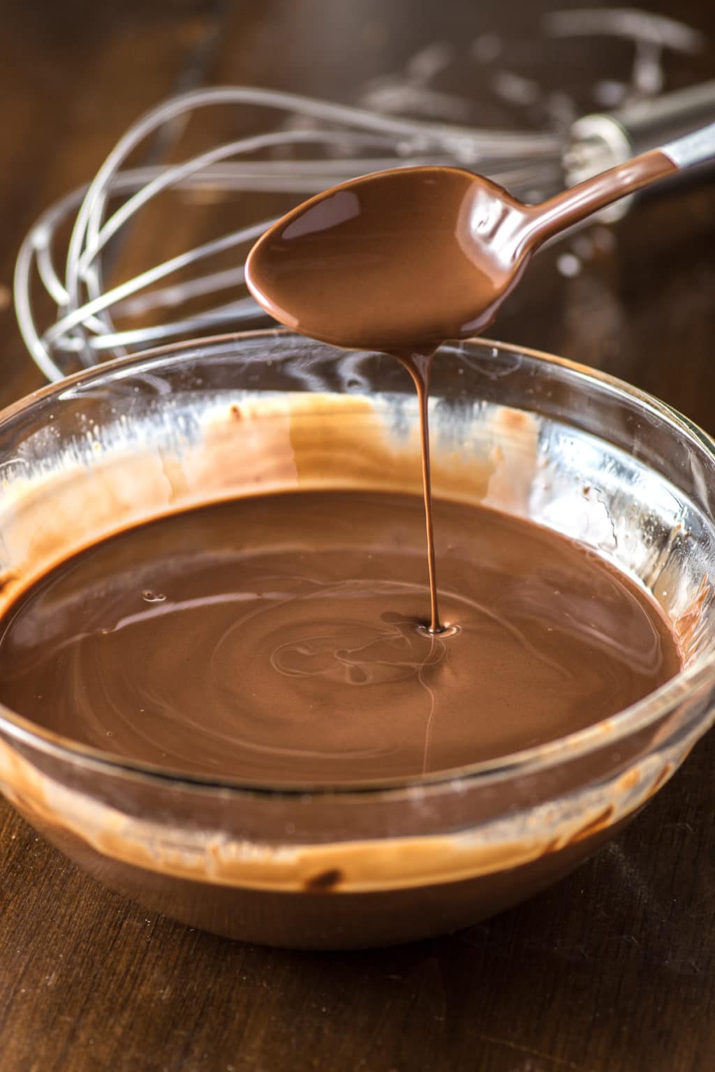 The clear glass mixing bowl is filled with molten chocolate with a teaspoon of melted chocolate held on top and dripped into the bowl. There is an egg beater in the background.