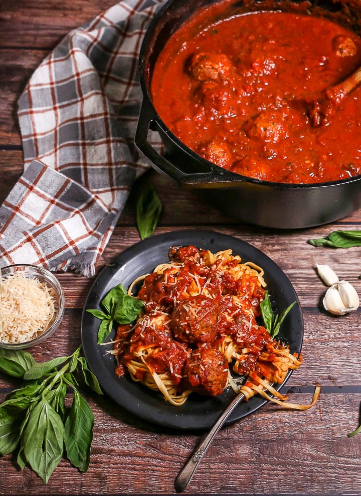 A portion of Sunday Sauce and Meatballs over fettuccine on a black plate garnished with fresh basil and a spinning fork in the pasta. The sauce pot is in the corner.