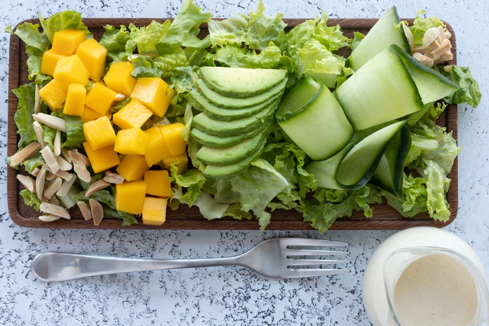 Aerial view of rectangular wooden plate with lettuce, mango, avocado and cucumber salad. Small glass jar with low acid salad dressing and fork on the bottom,