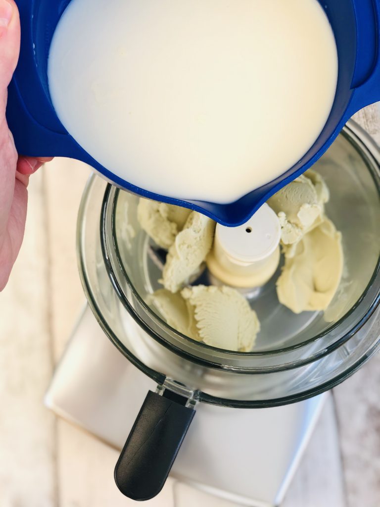 The magimix blender is filled with pistachio cream and milk is poured into a bowl, with a tub of ice cream beside the blender