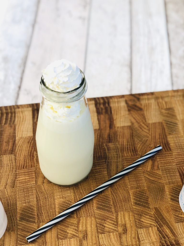 glass bottle filled with homemade pistachio ice cream milkshake with black and white striped reusable straw lying on a side table with milkshake sprayed on top of milkshake