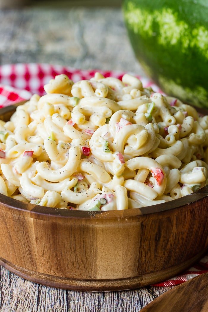 A wooden bowl with a red and white checkered napkin on a background full of creamy pasta salad.