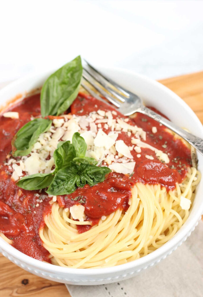 White bowl with spaghetti, marinara sauce, grated Parmesan cheese, fresh basil.
