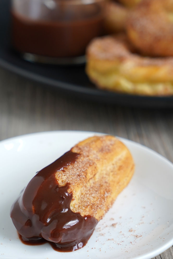 Baked churros dipped in chocolate sauce on a white plate with extra churros and sauce on the background.