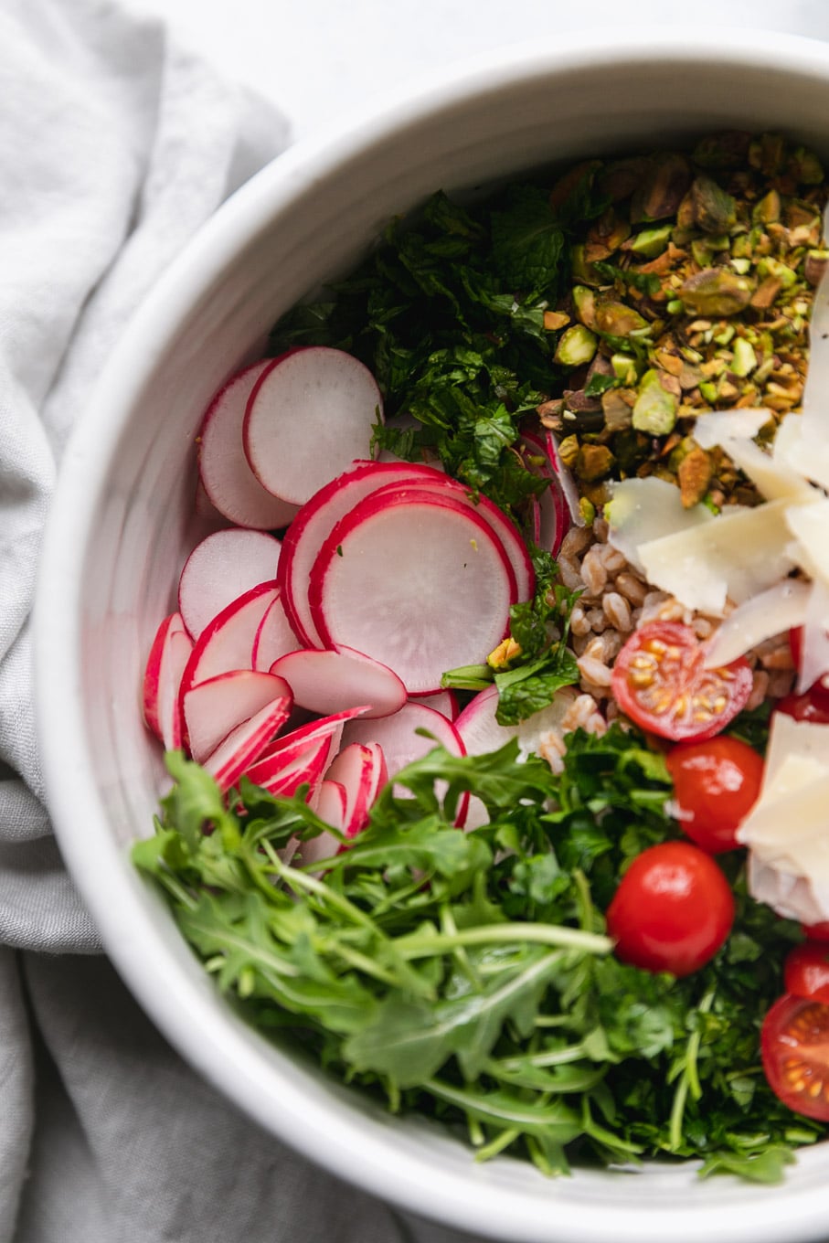 Aerial shot of a mixing bowl filled with arugula, topped with piles of radishes, grape tomatoes, grated parmesan and chopped pistachios