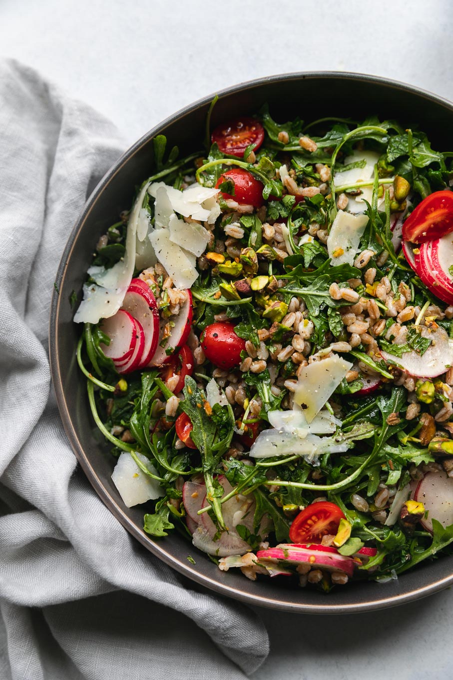 Aerial close-up of arugula farro salad topped with tomatoes, sliced ​​radish, shredded parmesan and chopped pistachios