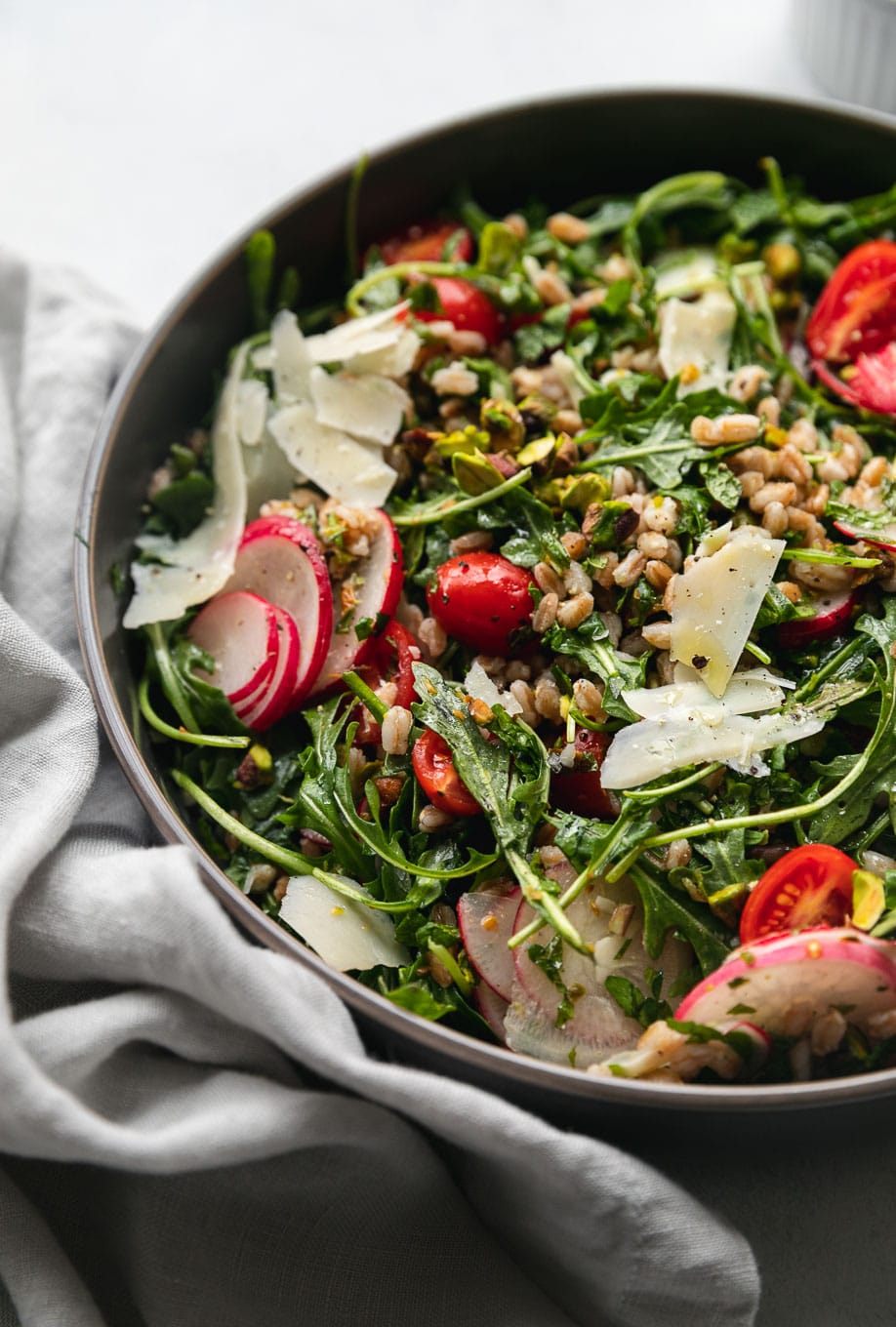 Close-up shot of arugula farro salad topped with tomatoes, sliced ​​radish, shredded parmesan and chopped pistachios