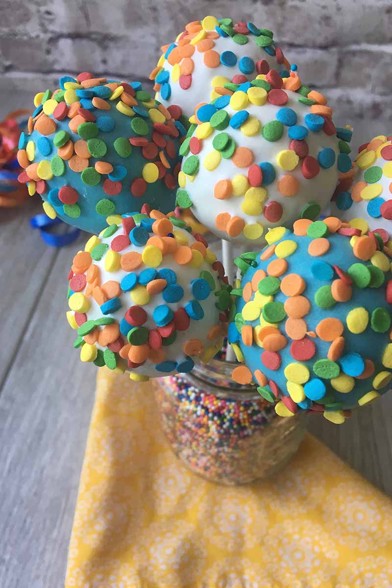 Vertical image of rainbow circle sprinkles covering assorted colored balls in a jar filled with more decorations.