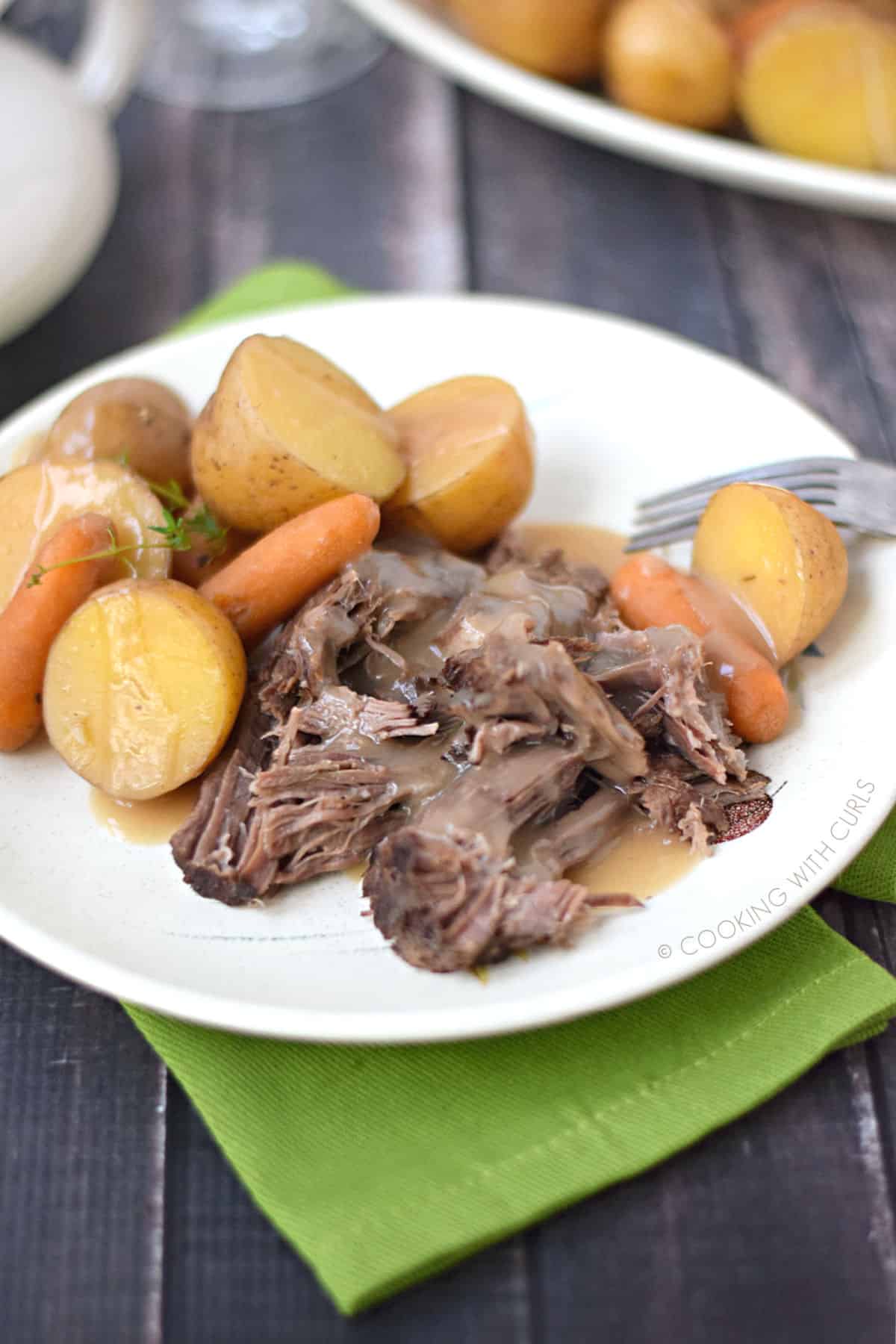 Roast pot with sliced ​​potatoes and young carrots covered in beef gravy on a white plate is placed on a green napkin.
