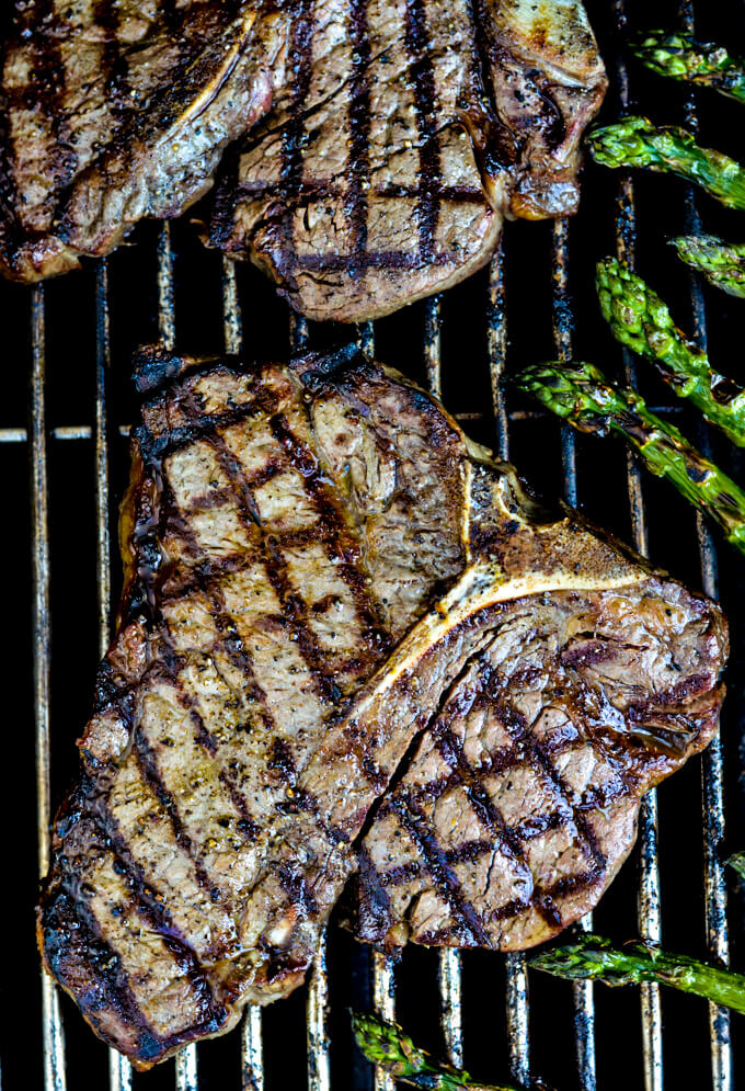 A steak on a white plate with a layer of melted blue cheese butter on top. A fork and a steak knife in the front and grilled asparagus on the plate next to the steak. Another plate with steak and asparagus in the back. A glass of red wine is next to the photo.