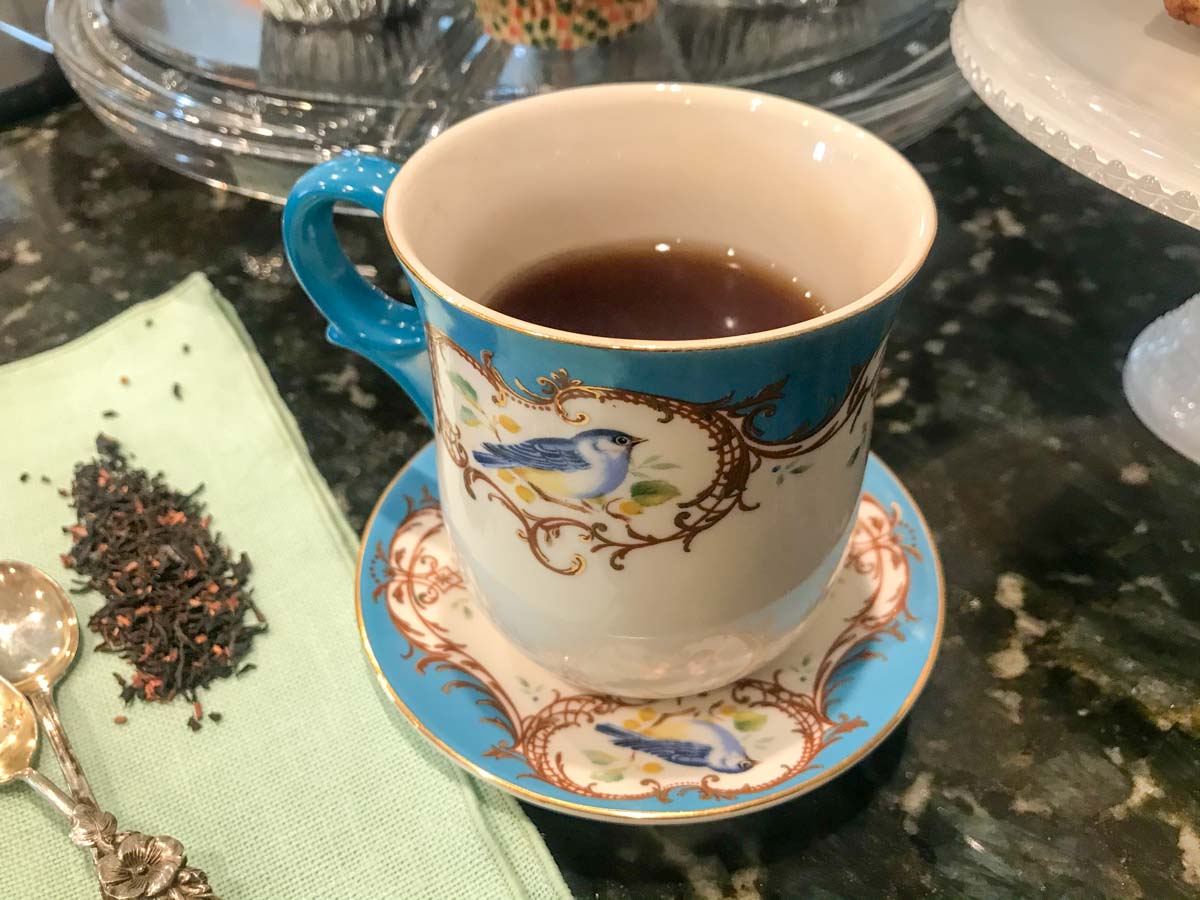 The image shows a cup of white green tea with a green bird on it, holding black tea. The cup rests on a black marble surface, next to a tea towel with teaspoons on it.