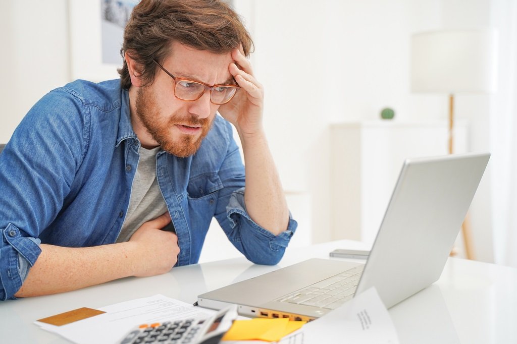 Worried man looking at computer screen with utility bill on desk.