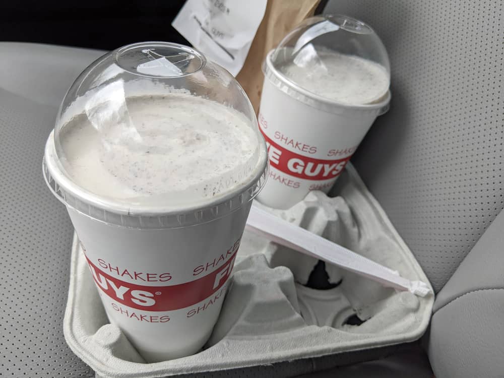 View of two milkeshakes and an order of fries from Five Guys sitting in the passenger seat of a car