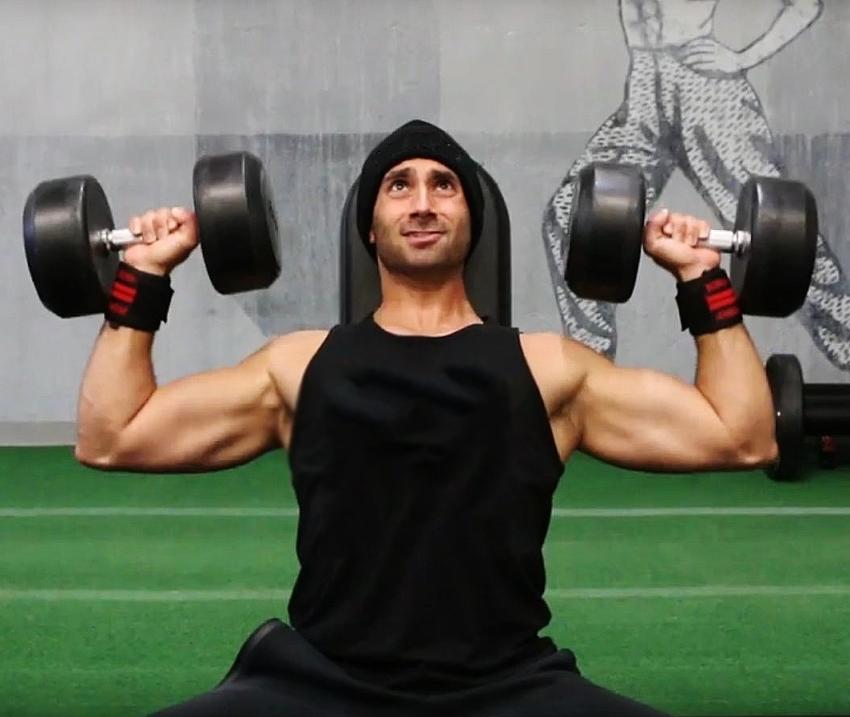 Dom Mazzetti doing shoulder presses while sitting on a bench