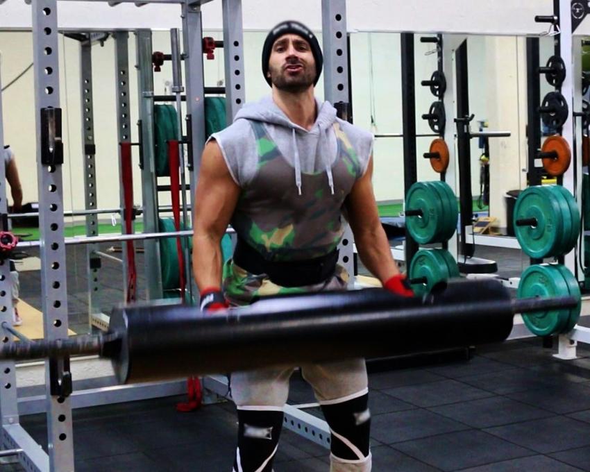 Dom Mazzetti lifts a black barbell while saying something ironic in front of the camera