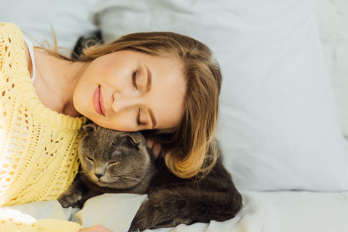 Woman curled up with her eyes closed with a sleepy cat on the bed