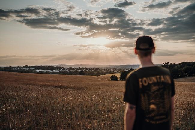 A young man stands with his back to the camera, looking at the sun shining its bright rays across the earth, through low clouds. Jesus came to earth and was taught by many people, including God and Mary and Joseph.