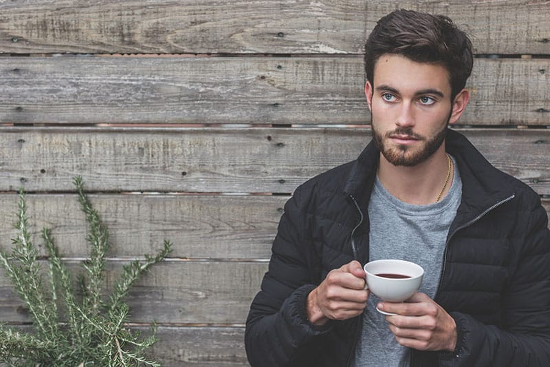 man holding coffee cup leaning against wooden wall